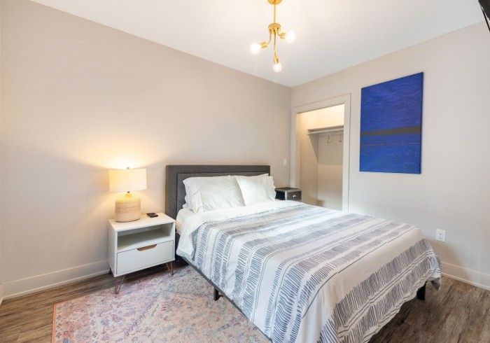 Bedroom with bed, nightstand, art, and closet. Pale walls, wood floors, decorative rug.