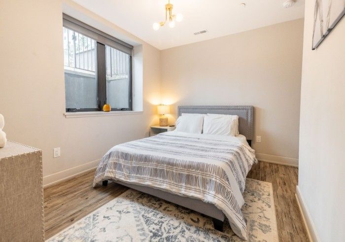 Bedroom with double bed, small rug, window, neutral walls, bedside table with lamp, and a modern light fixture.