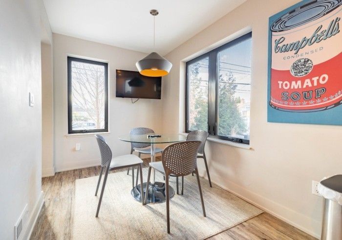 Dining area with glass table, four chairs, a Campbell's Soup can art print, windows, and pendant light.