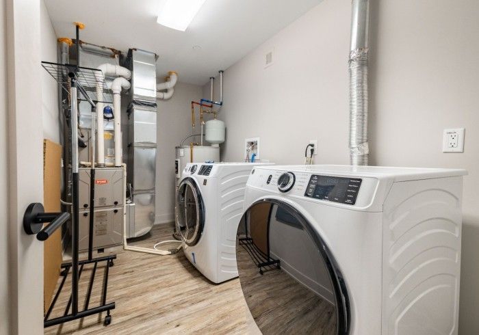 Laundry room with white washer and dryer, appliances, and vents. Light wood flooring.
