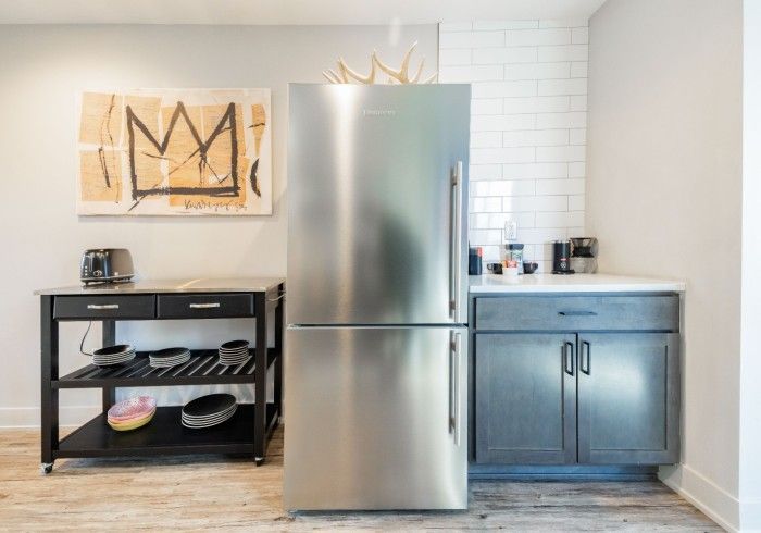 Kitchenette with a stainless steel refrigerator, black cart, and gray cabinets. Art on the wall.