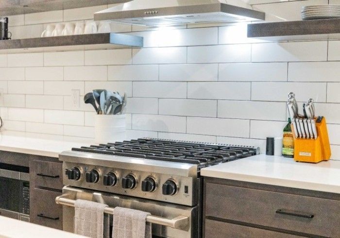 Stainless steel stove in a modern kitchen with white subway tile backsplash, wooden shelves, and gray cabinets.