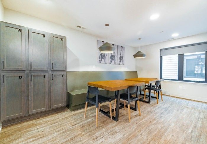 Locker-lined room with wood tables, black chairs, green bench seating, and two pendant lights. A window is on the right.