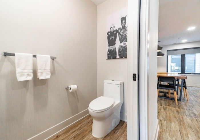 Bathroom with a white toilet, towels, and a framed black and white photo.  Doorway leads to a dining area.