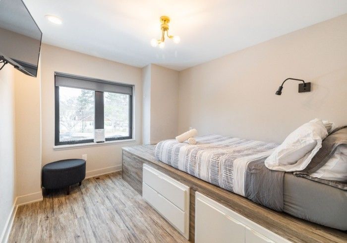 Bedroom with a bed on a raised platform, window, TV, and black ottoman. Light wood flooring and neutral walls.