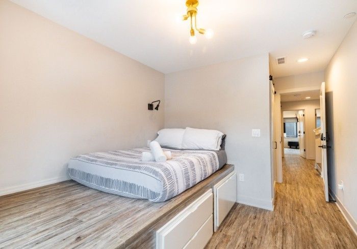 Bedroom with bed on platform; drawers below. Light-colored walls and wood-look flooring. Hallway visible.