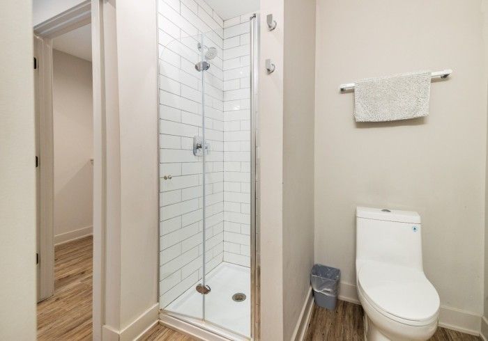 Bathroom with white-tiled shower, glass door, toilet, towel rack, and trash can. Light-colored walls and flooring.