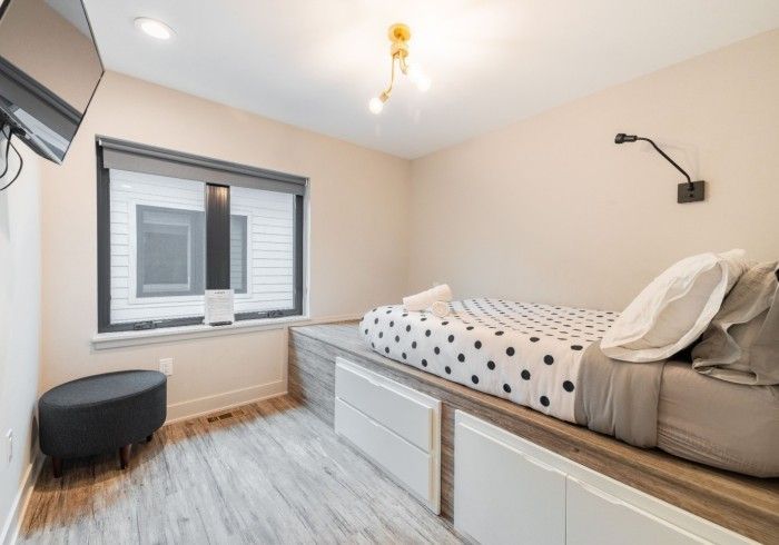 Bedroom with a built-in bed frame, window with dark frame, black polka dot bedding, a black ottoman, and light-colored walls.