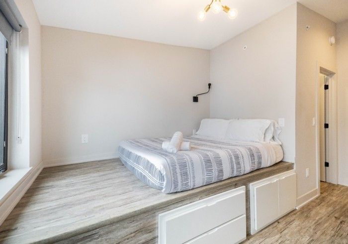 Bedroom with a bed on a raised platform, white walls, large window, and light wooden flooring.