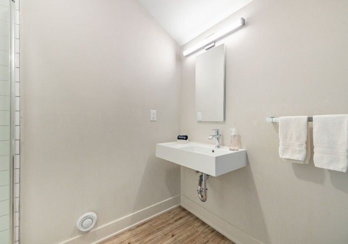 Small, modern bathroom with white floating sink, mirror, and towel rack. Off-white walls, wood-look floor.