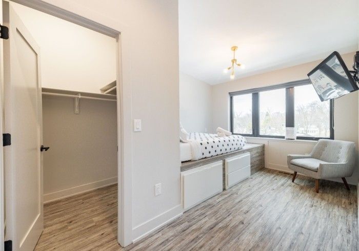 Bedroom with built-in bed platform, chair, and closet. Light wood floor, white walls. TV on wall.