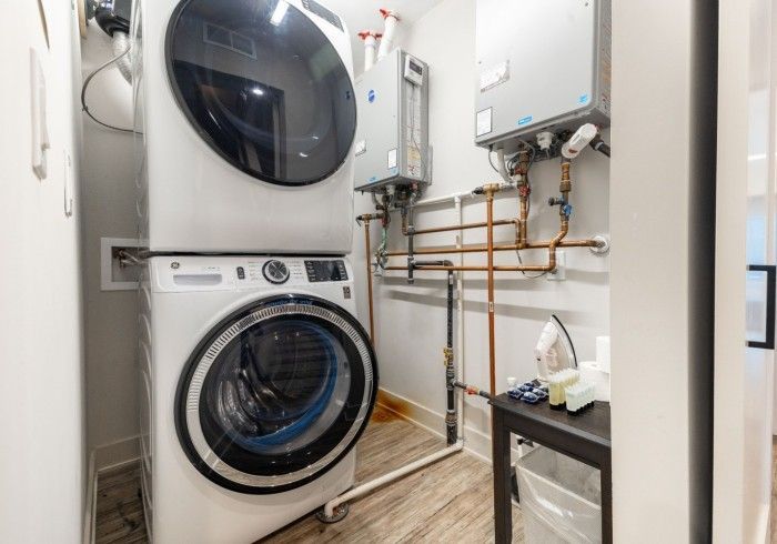 Laundry room with stacked washer/dryer, water heater, copper pipes, small black table, and iron.
