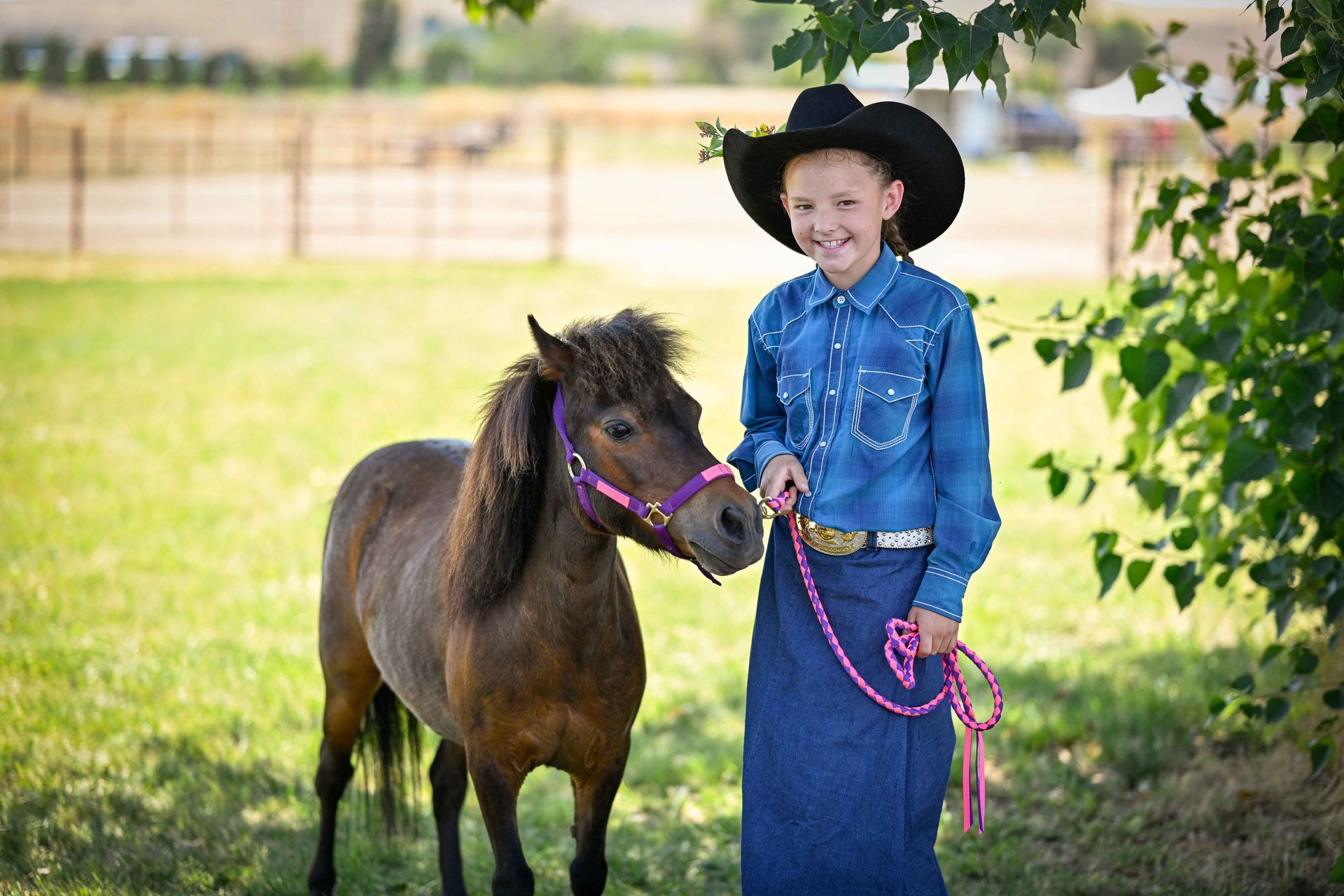 A young girl in a cowboy hat is standing next to a pony in a field.