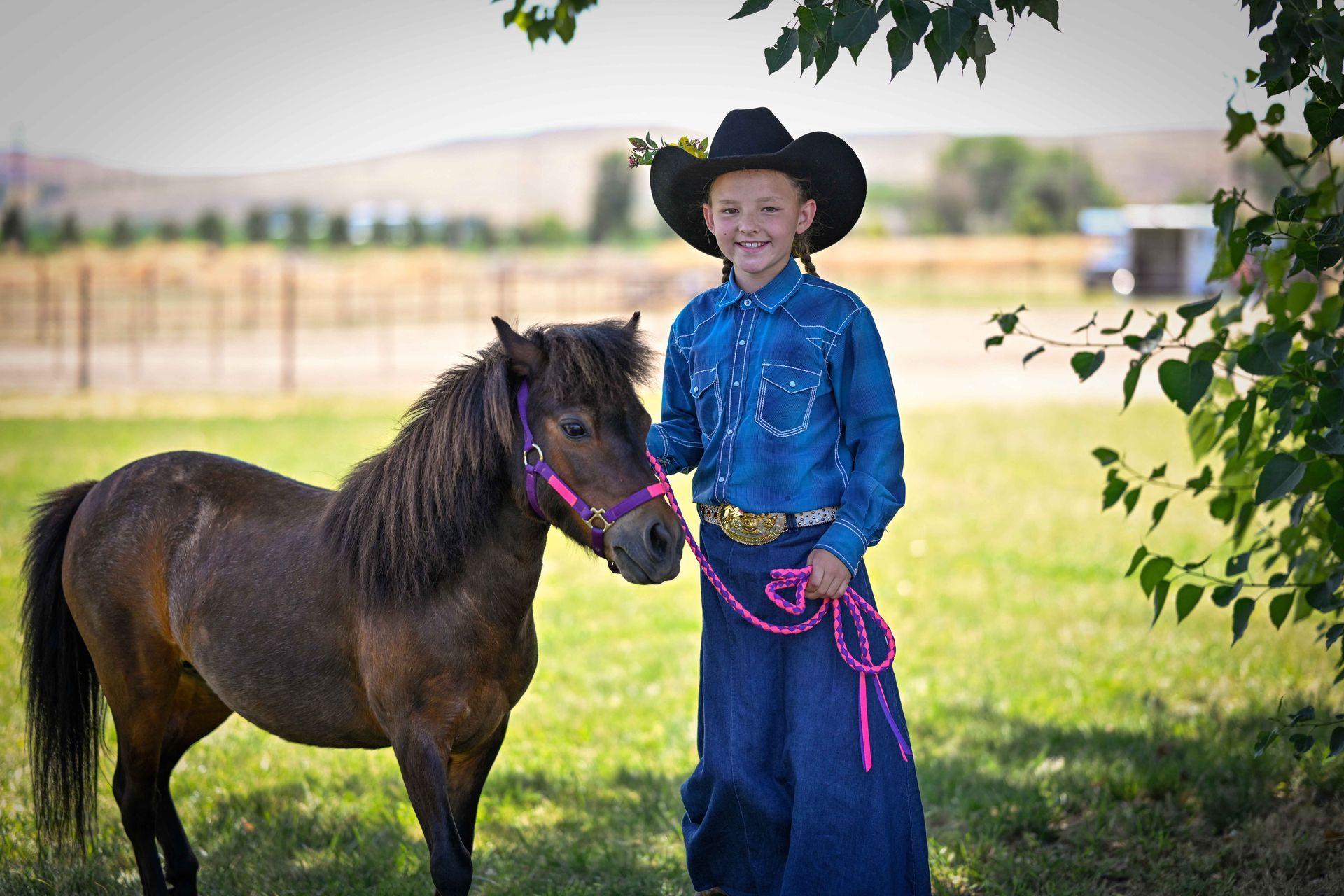 A young boy in a cowboy hat is standing next to a small brown horse.