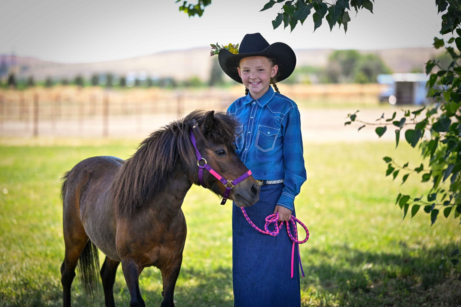 A young girl in a cowboy hat is standing next to a small brown horse.