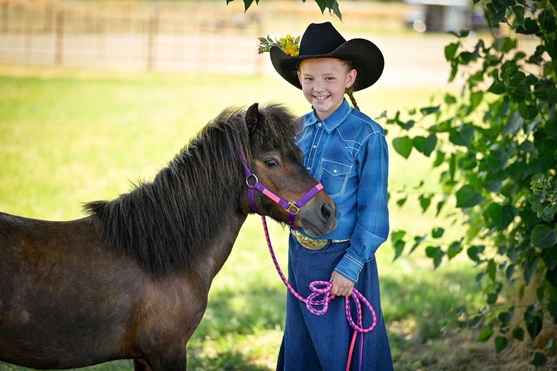 A young girl in a cowboy hat is standing next to a small brown horse.