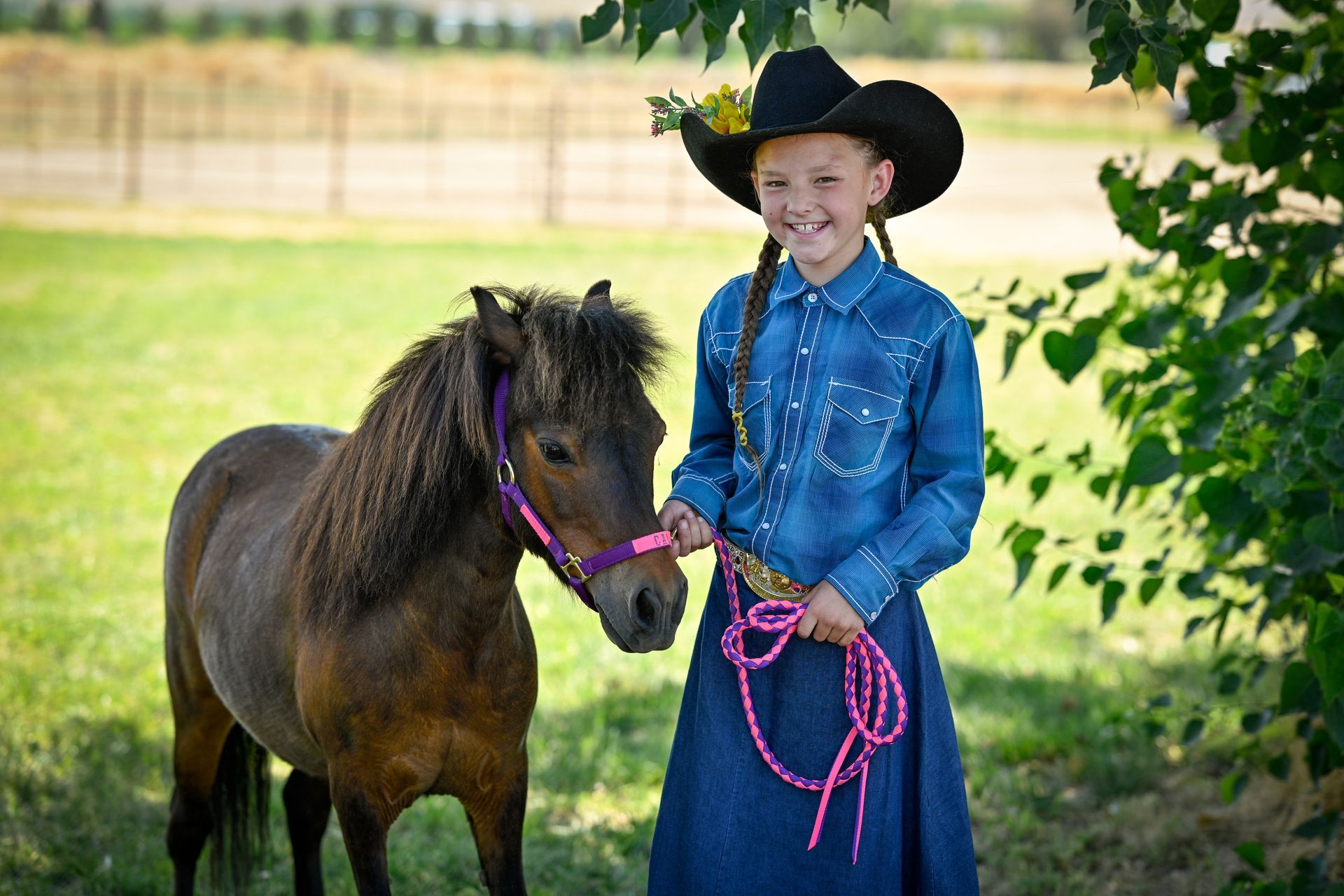 A young girl in a cowboy hat is standing next to a small brown horse.