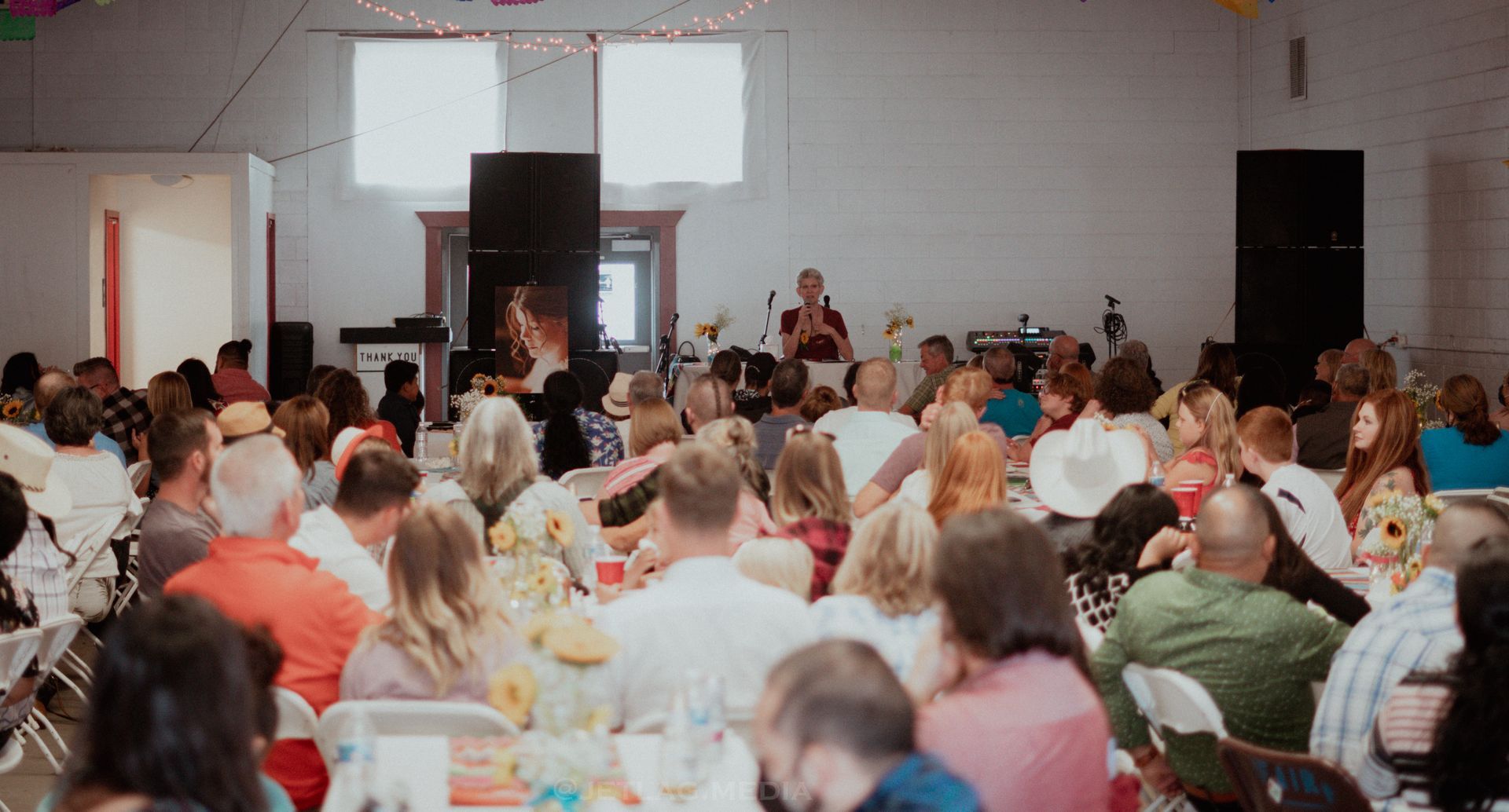 Large crowd seated at tables in a brightly lit hall, listening to a speaker on a stage.