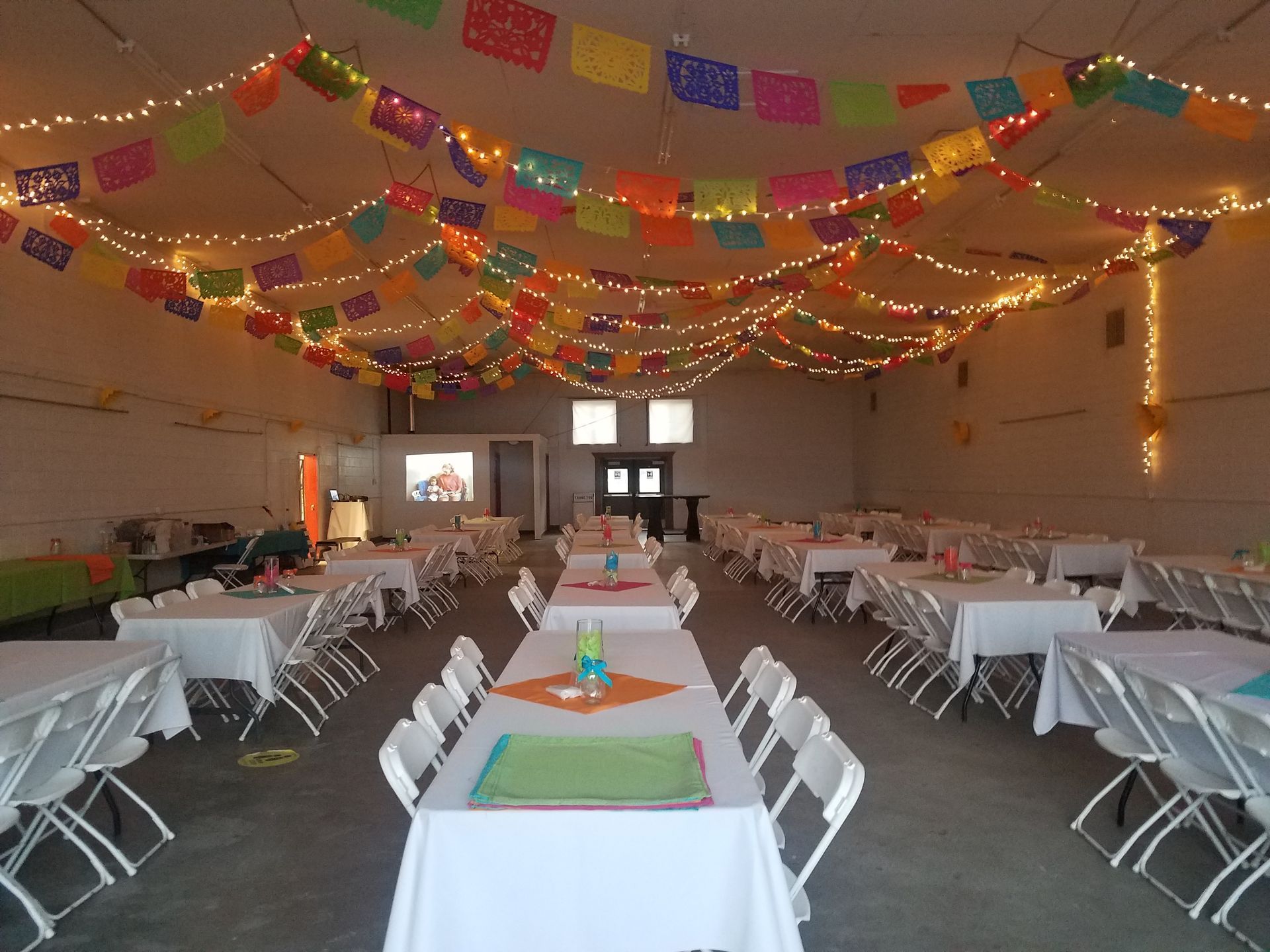 Party hall with white tables, chairs, colorful flags, string lights, and a projector screen.