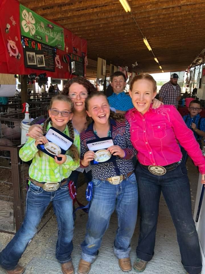 Group of girls and women, some wearing cowboy hats and holding ribbons, smiling under a covered area.