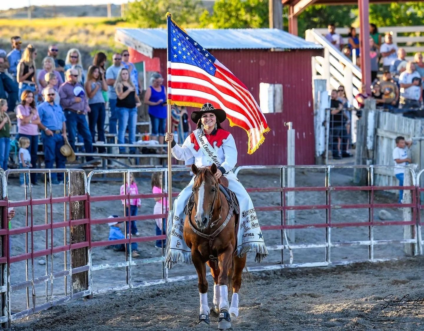 Rodeo queen on a horse waves American flag, red, white, and blue, in a rodeo arena with cheering crowd.