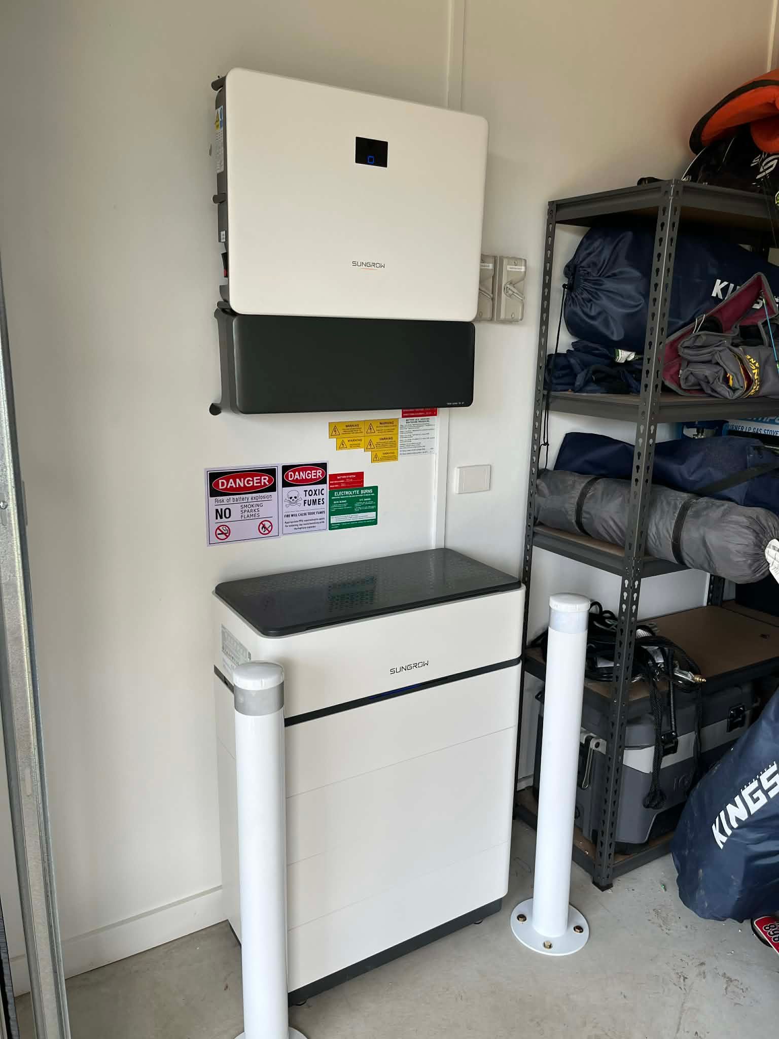 White solar inverter and battery on a wall, with safety signs and a metal shelving unit nearby.