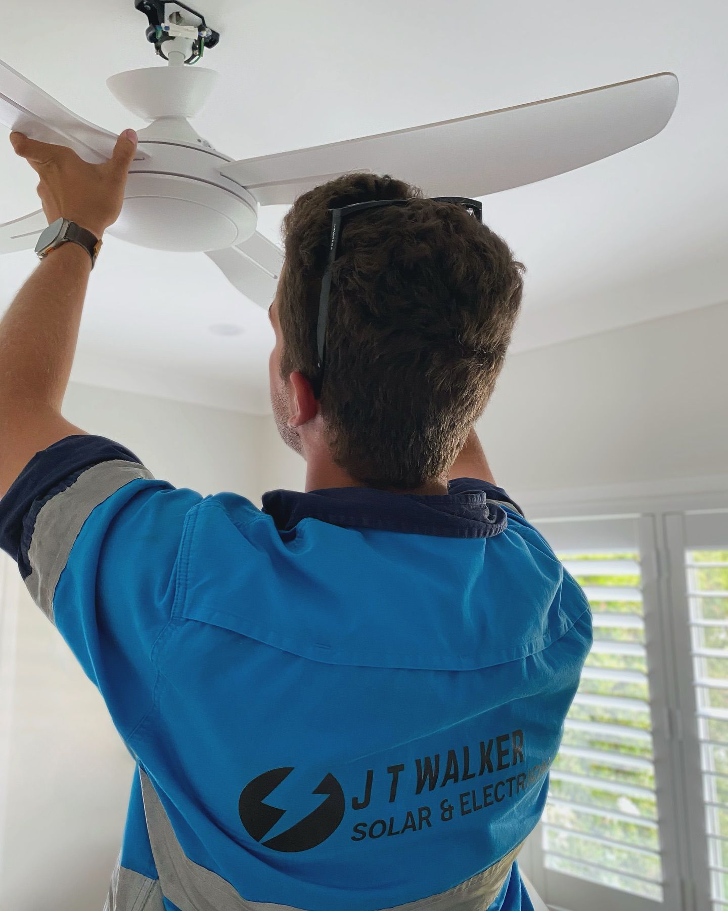 Man in blue shirt installs ceiling fan; interior setting.