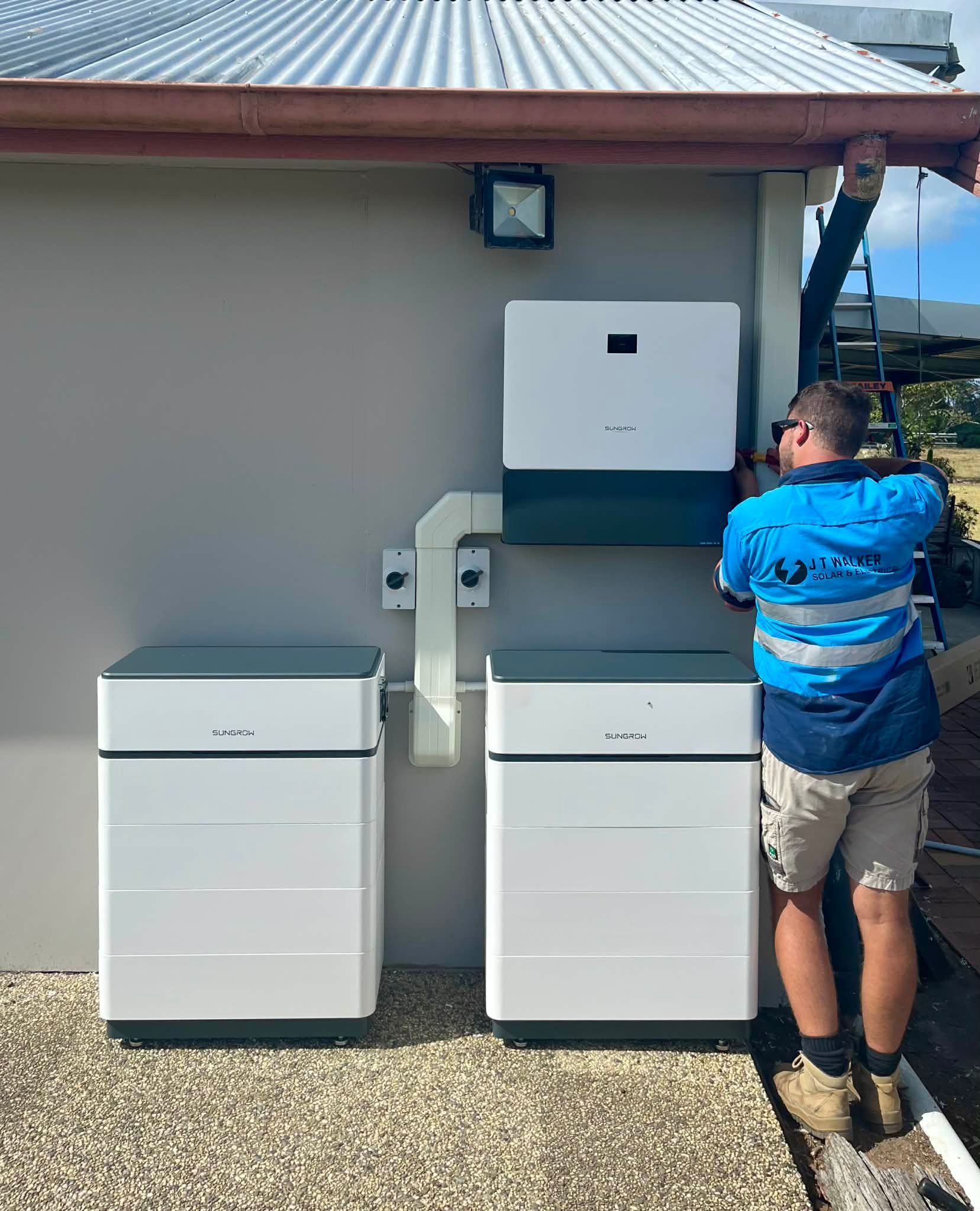 Electrician installing solar batteries on a building. White boxes, gray wall, blue and white shirt.