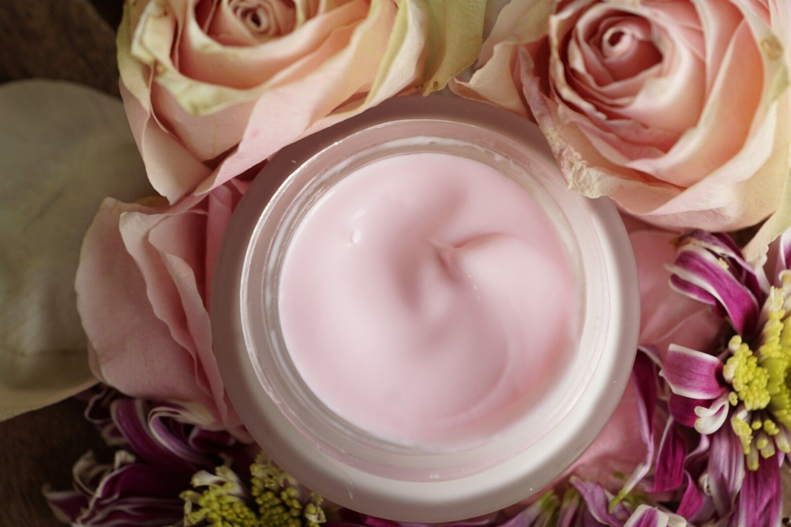 A jar of pink cream is surrounded by pink roses.