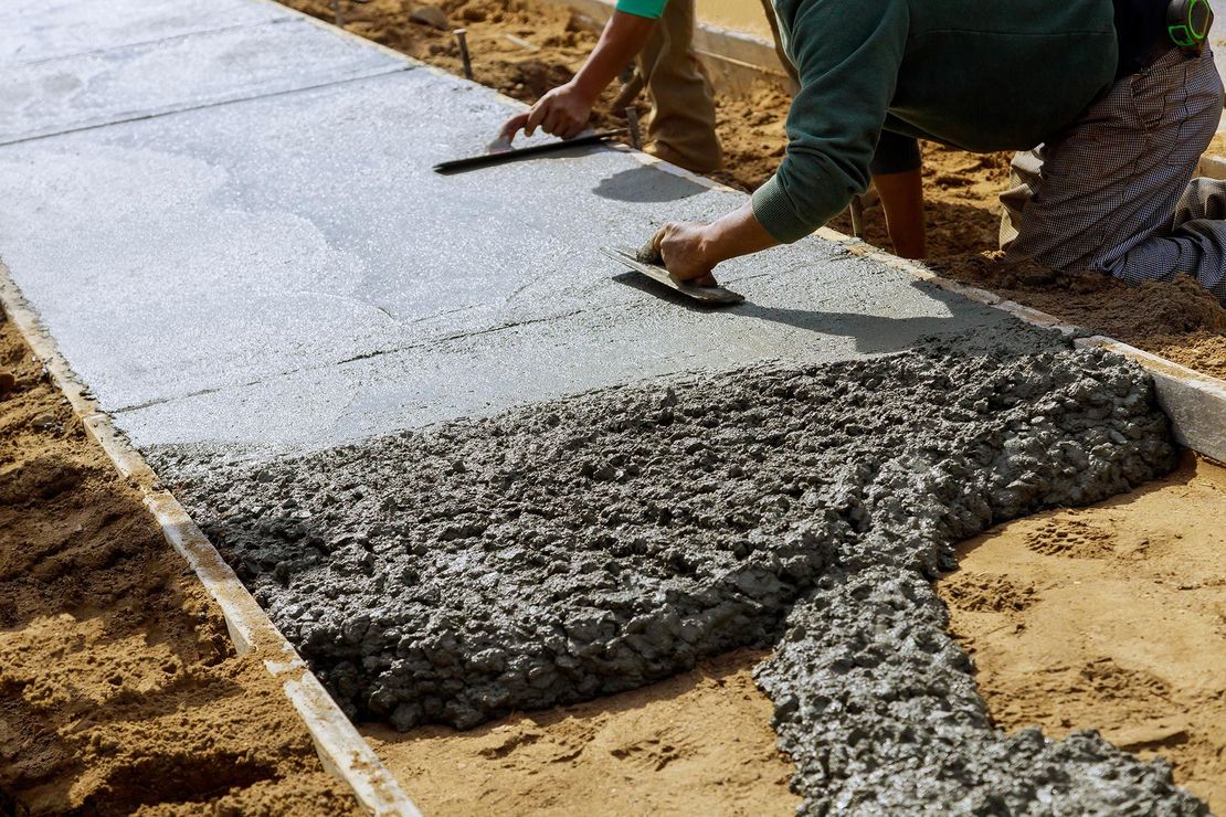 A construction worker smoothing wet concrete with a trowel to create a sidewalk. The concrete is grey, and the surrounding ground is sand.