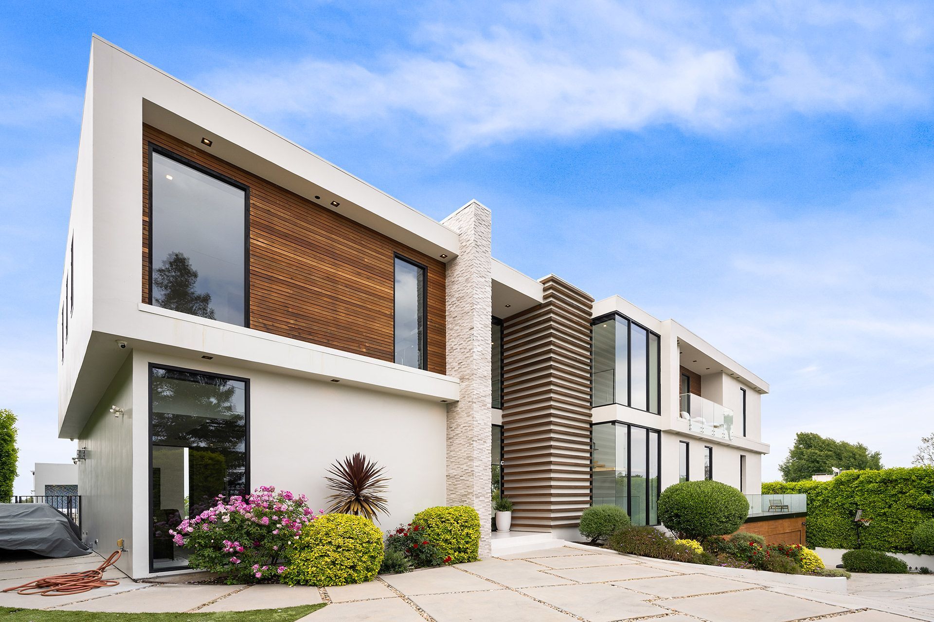 Modern two-story house with white walls, wooden paneling, and large windows. Landscaped yard with shrubs and flowers; blue sky overhead.