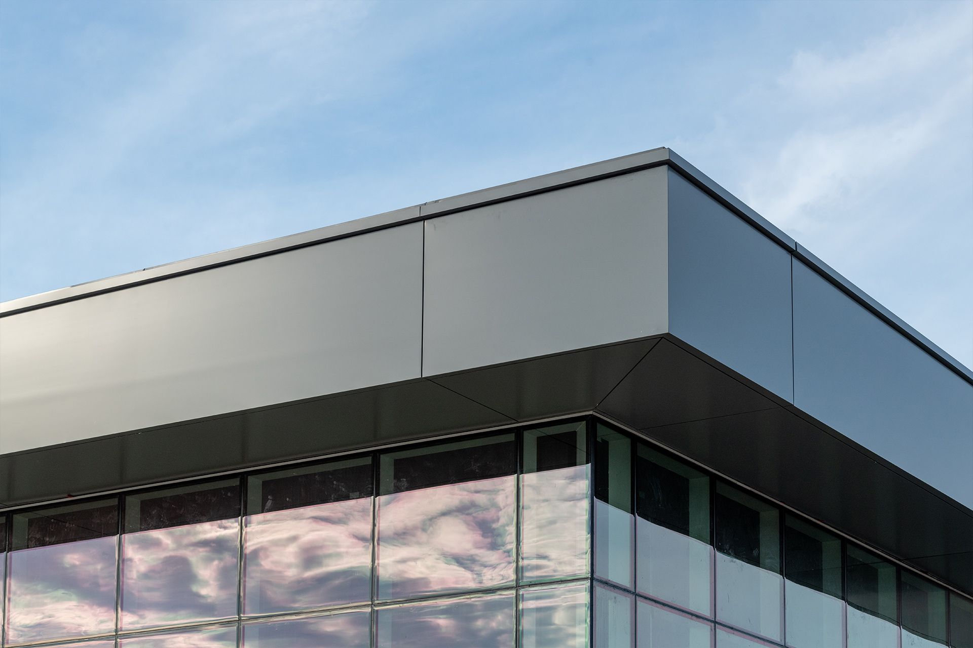 Corner of a modern building with glass windows reflecting the sky. It features a gray, angular roof and clear blue sky above.