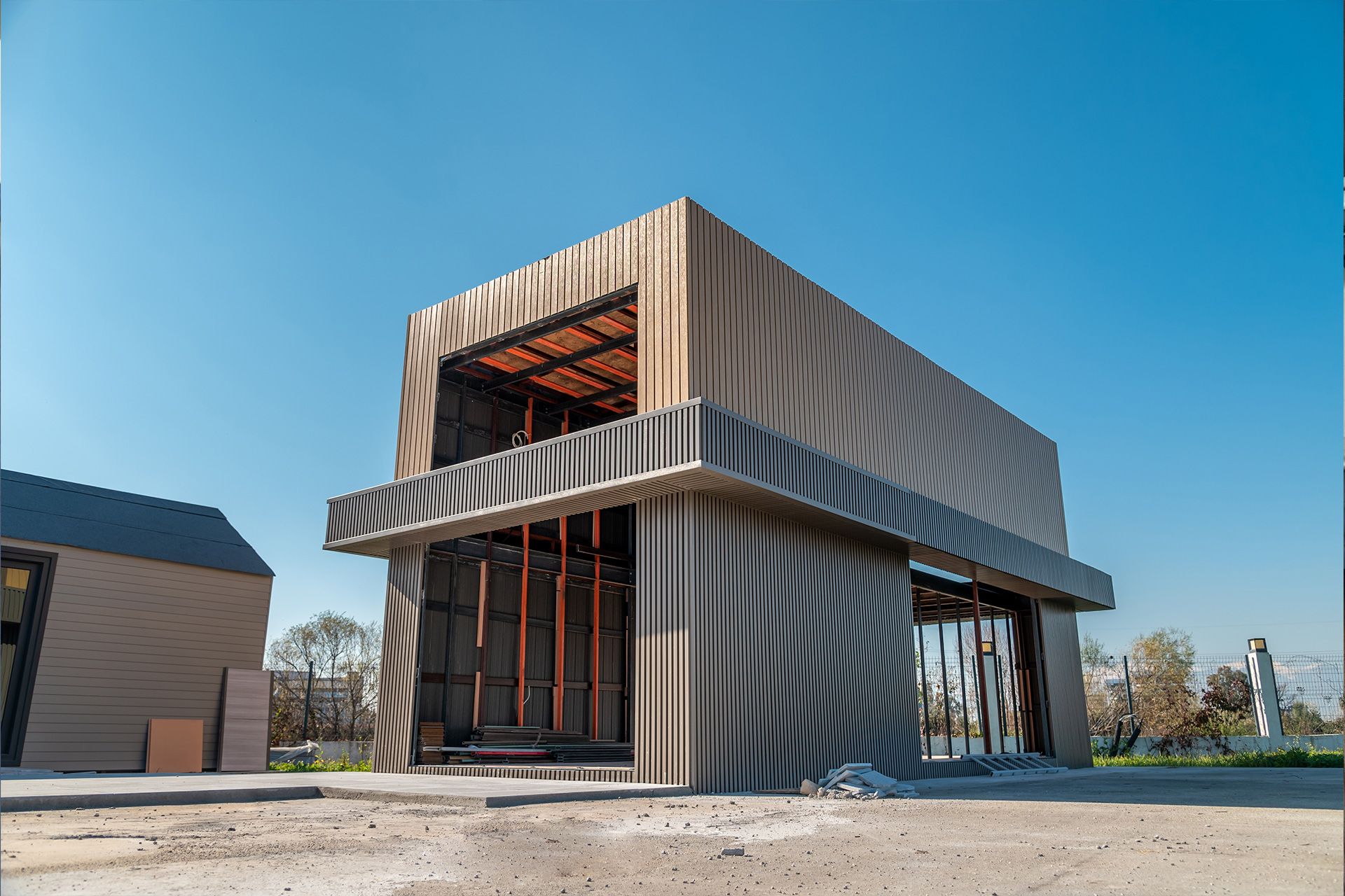 Modern building under construction with ridged metallic siding, open sides, and a concrete overhang, set against a clear blue sky.