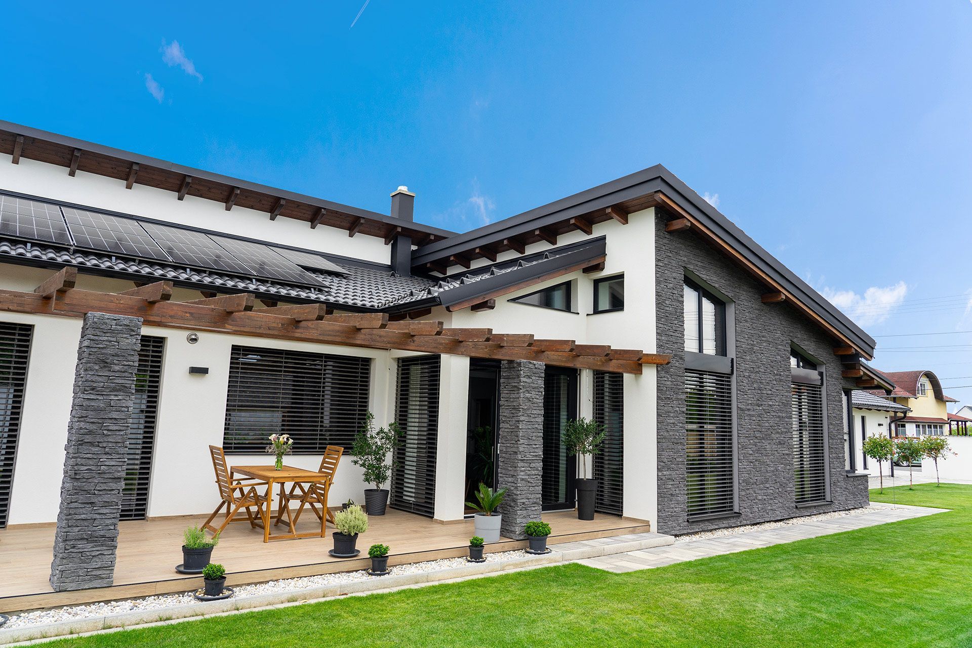 Modern house exterior with gray brick and white walls, wooden pergola, and patio furniture on a green lawn under a blue sky.