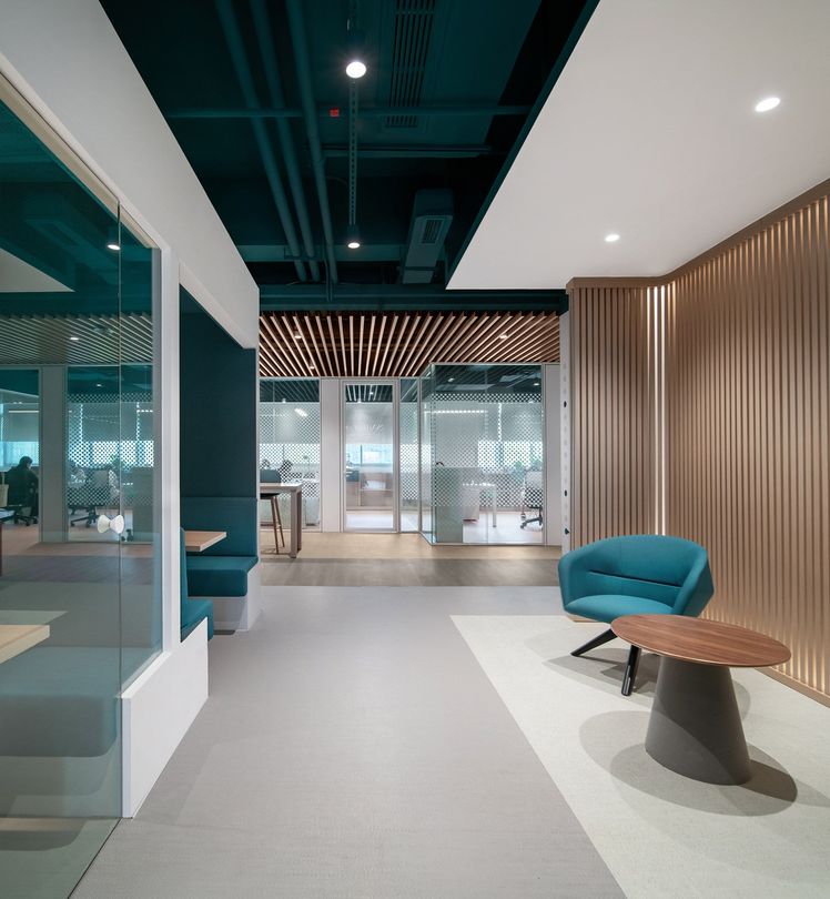 Interior office space with teal and beige accents; seating area with blue chair and small round table, vertical wooden paneling on the right, glass-walled office spaces in the background.