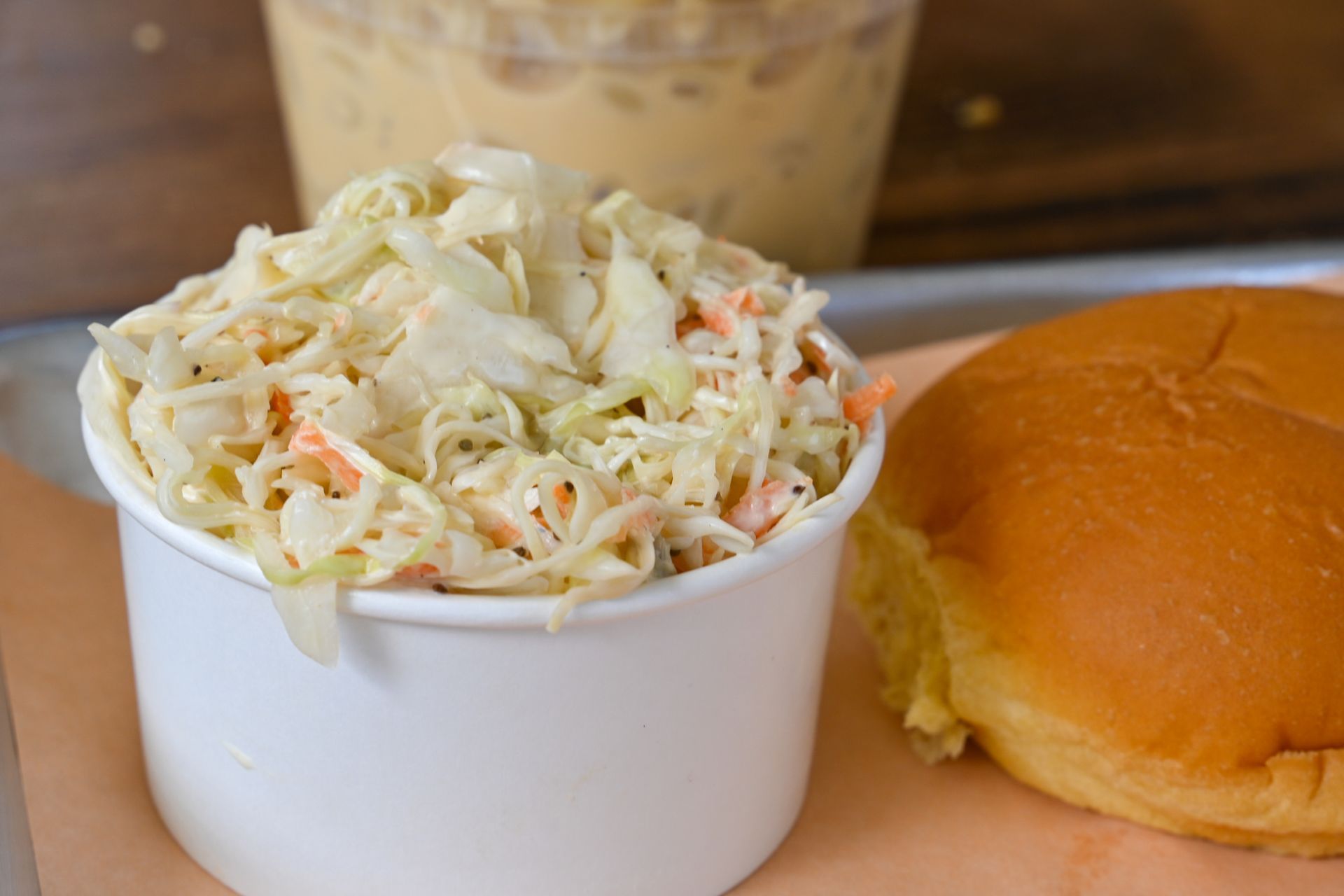 Coleslaw in a white container, with a bun on a tray. Creamy sauce in background.