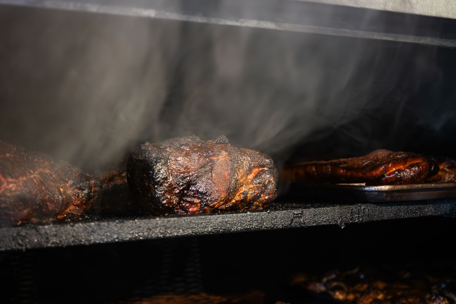 Smoked meat inside a smoker, with steam rising. Dark brown crust, metal grates, and dim light.