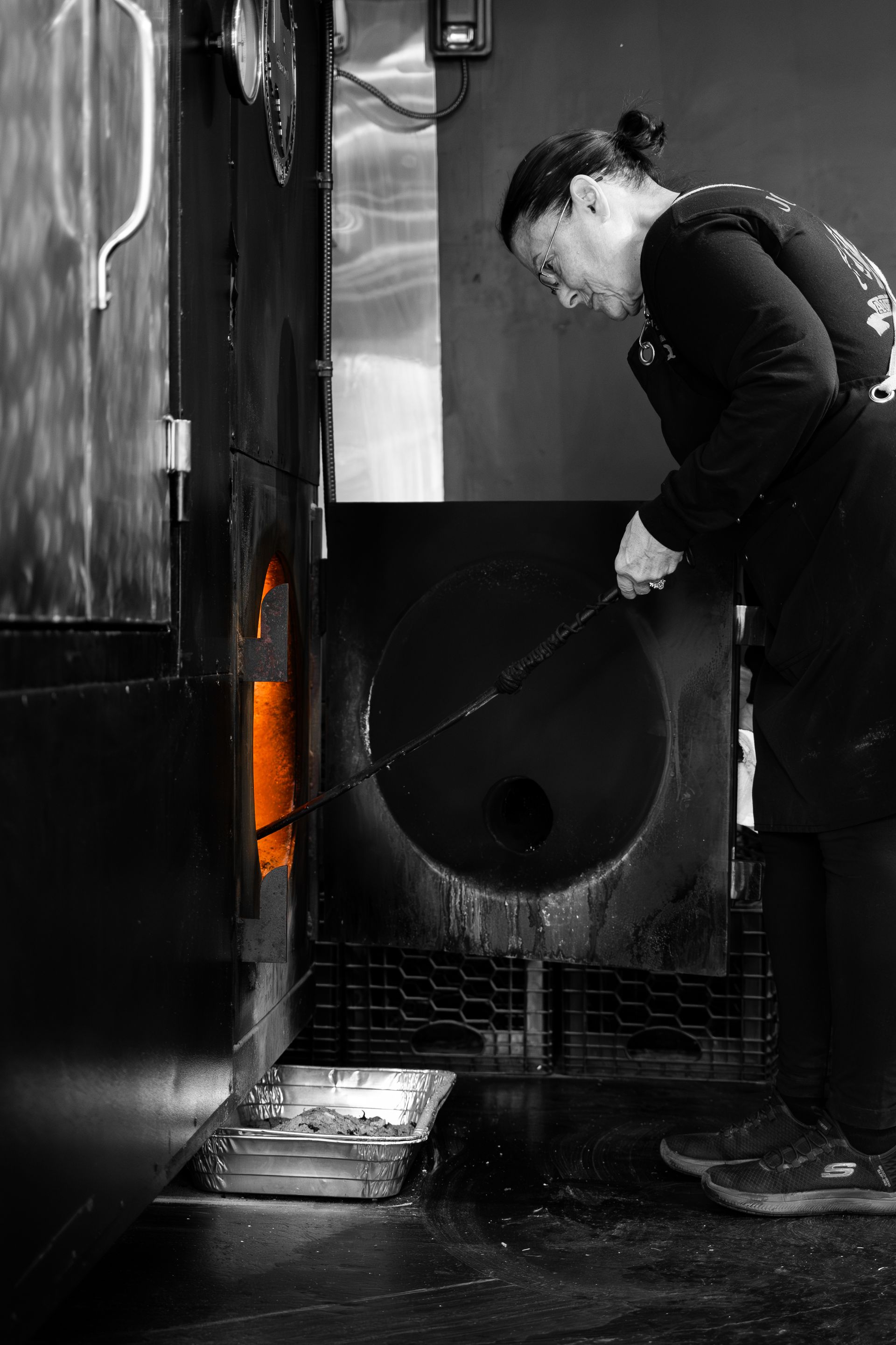 Woman working with molten glass in a furnace, wearing apron and looking down. The interior glows orange.