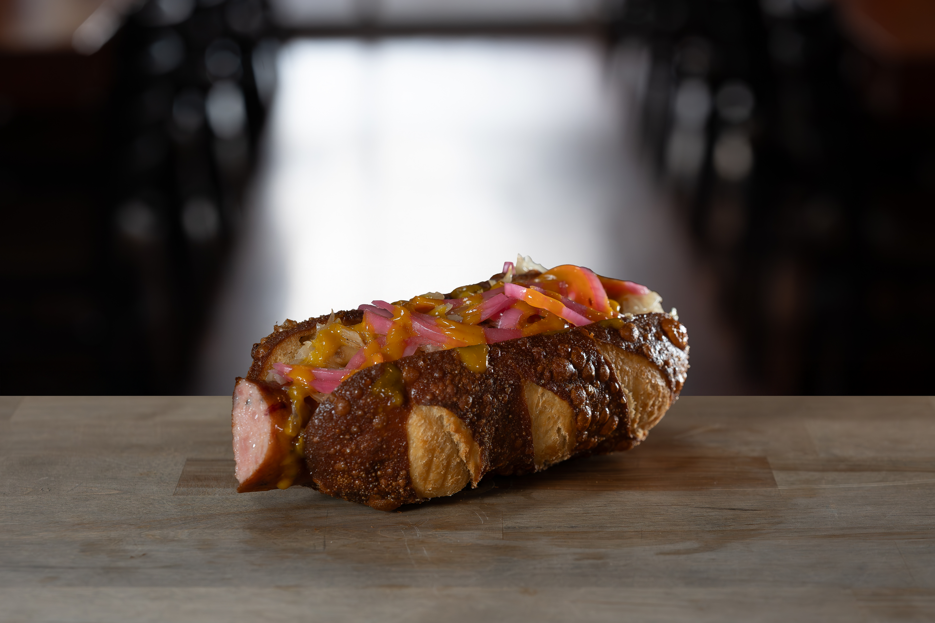 Hot dog on pretzel bun topped with pink onions and yellow cheese. Wooden surface, blurred restaurant background.