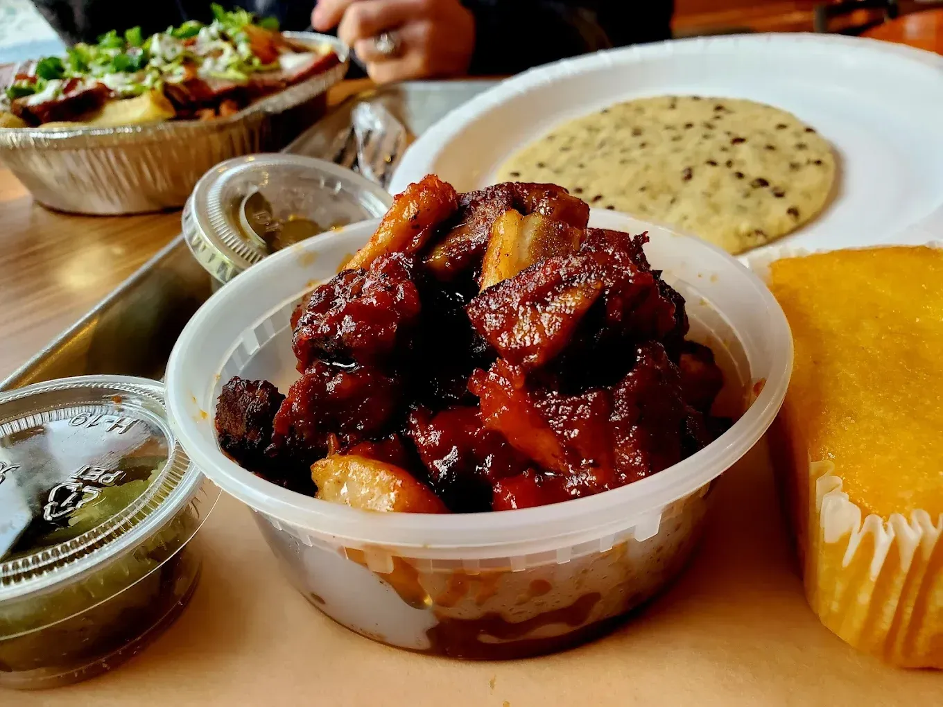 Barbecue ribs in a plastic container, corn bread, and a tortilla on a table.