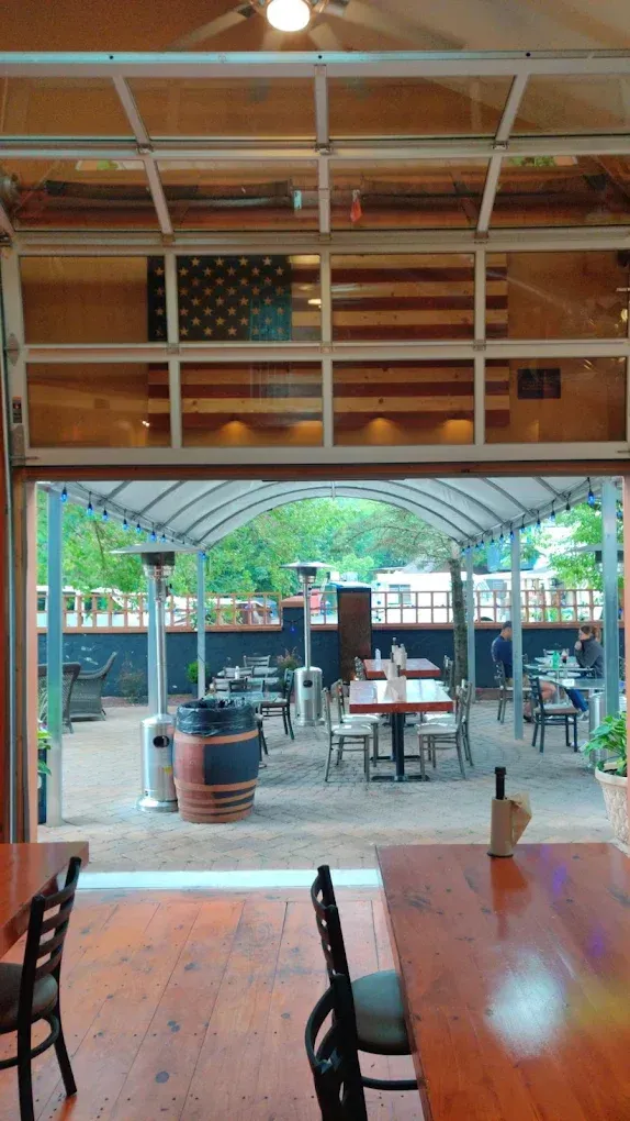 Restaurant interior with an outdoor patio. American flag visible through the open garage door. Tables and chairs on patio.