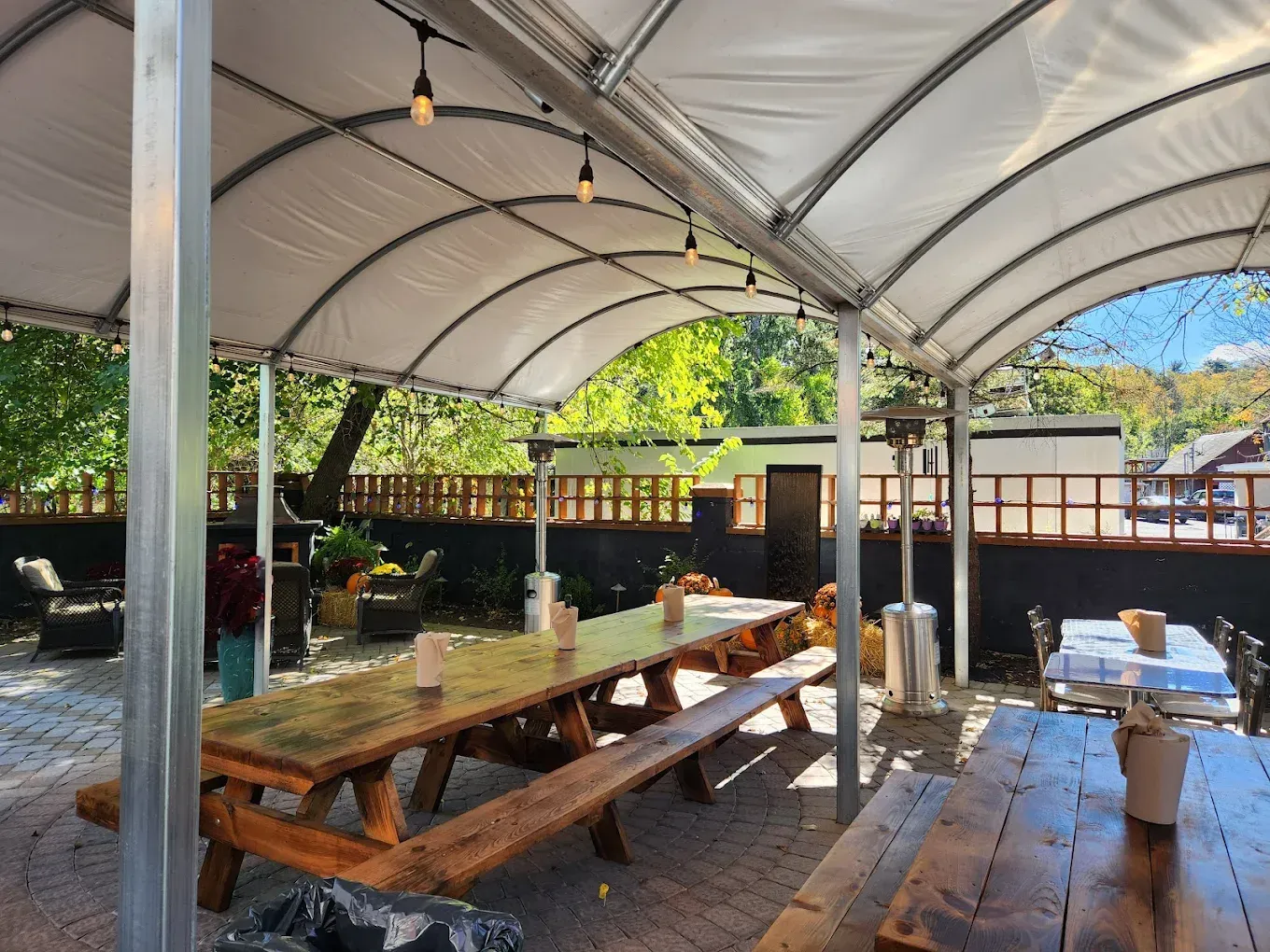 Outdoor dining area with picnic tables under a tent canopy, trees and buildings in background.