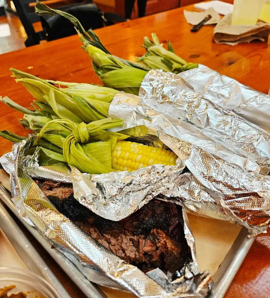 Corn on the cob wrapped in foil, next to smoked meat on a tray.