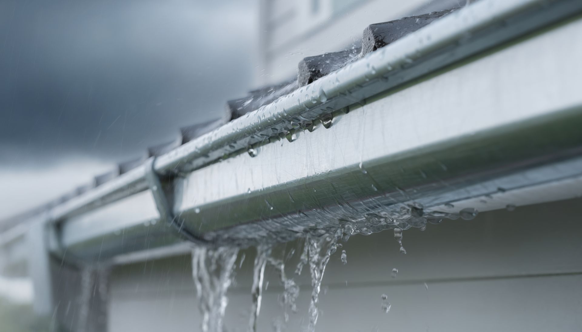 Rainwater overflowing from a light-colored metal gutter against a dark, stormy sky.