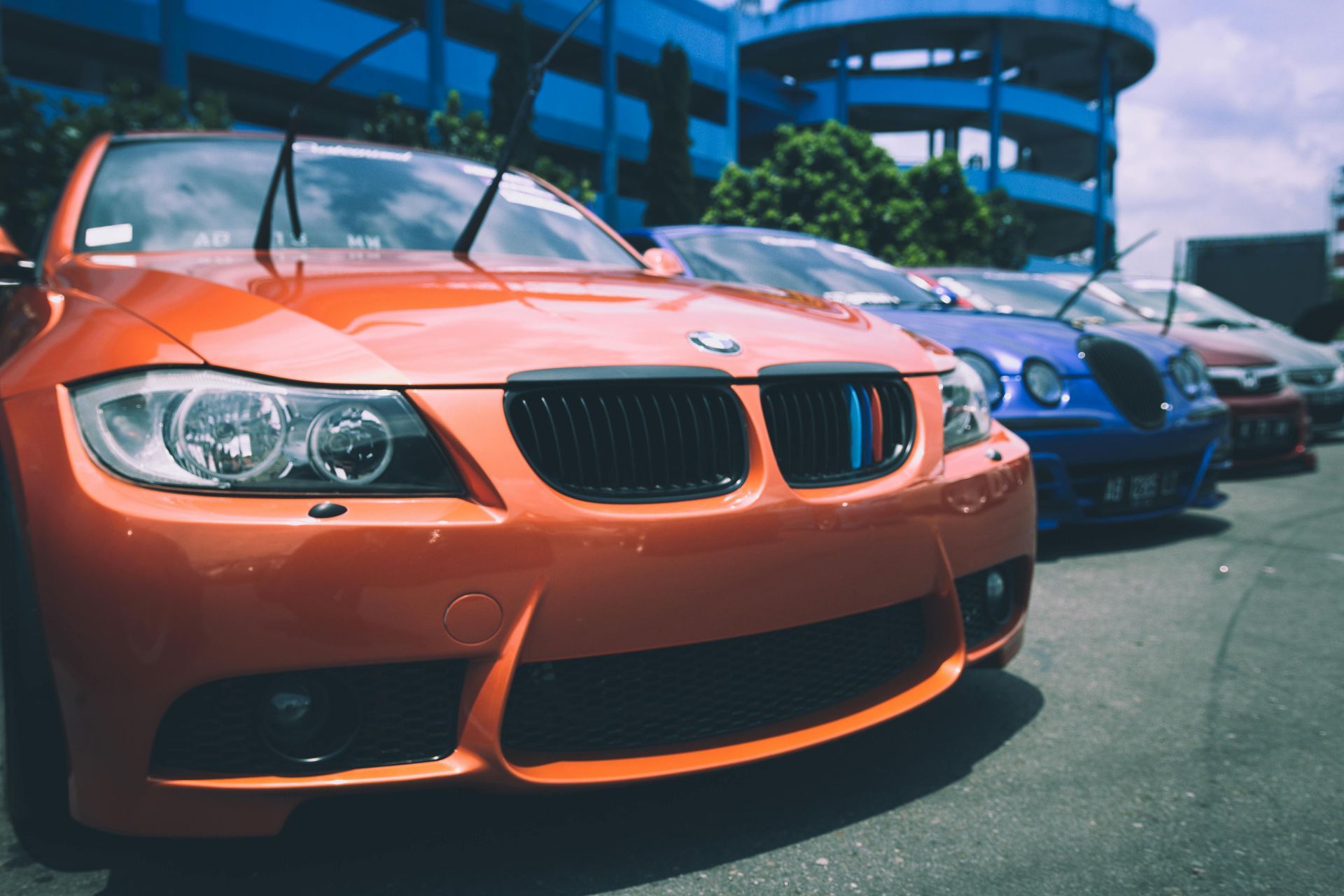 Orange BMW sports car parked in front of a blue parking structure, other cars in the background.