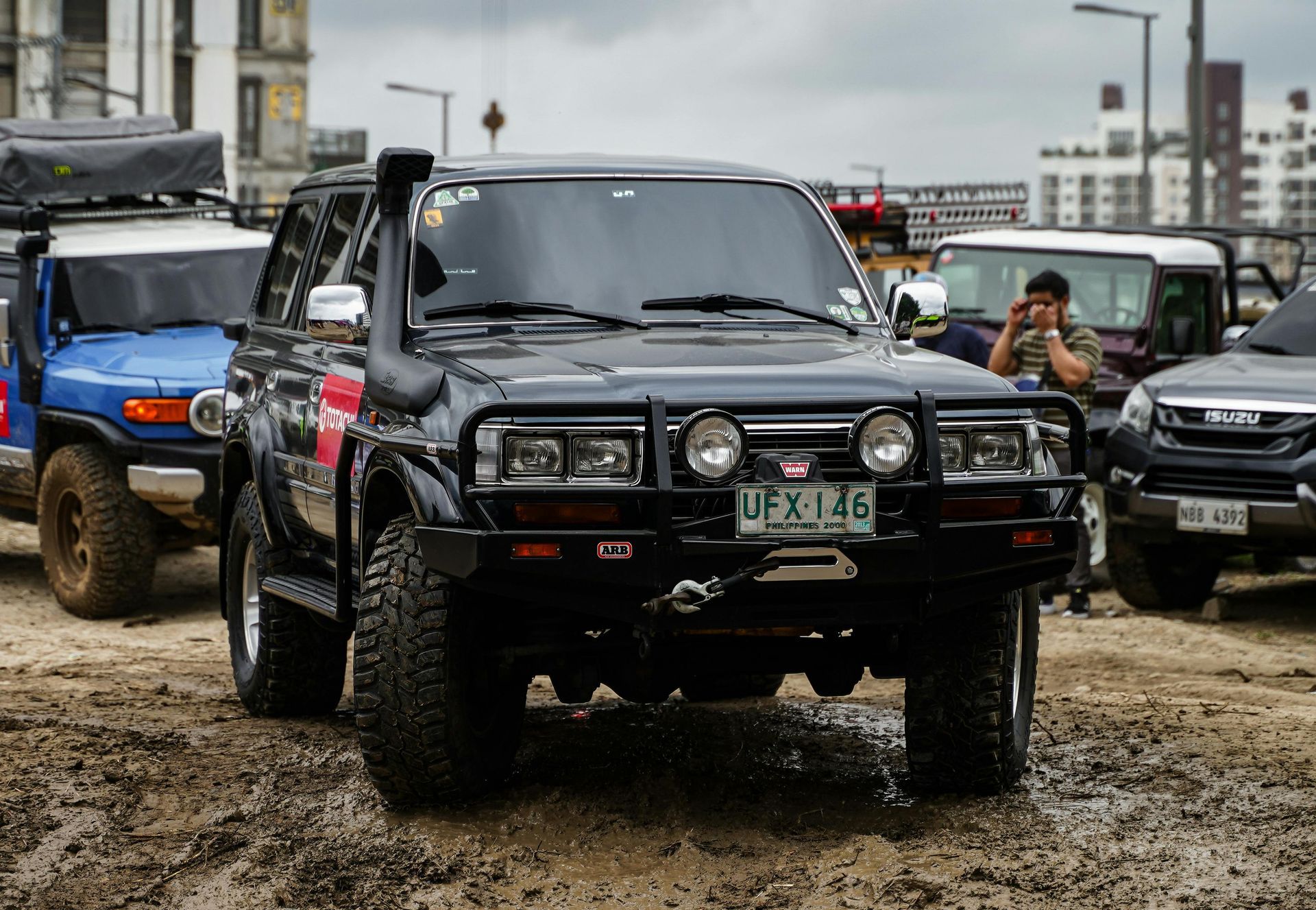 Dark off-road vehicle with snorkel, bull bar, and spotlights in a muddy outdoor setting with other modified vehicles.