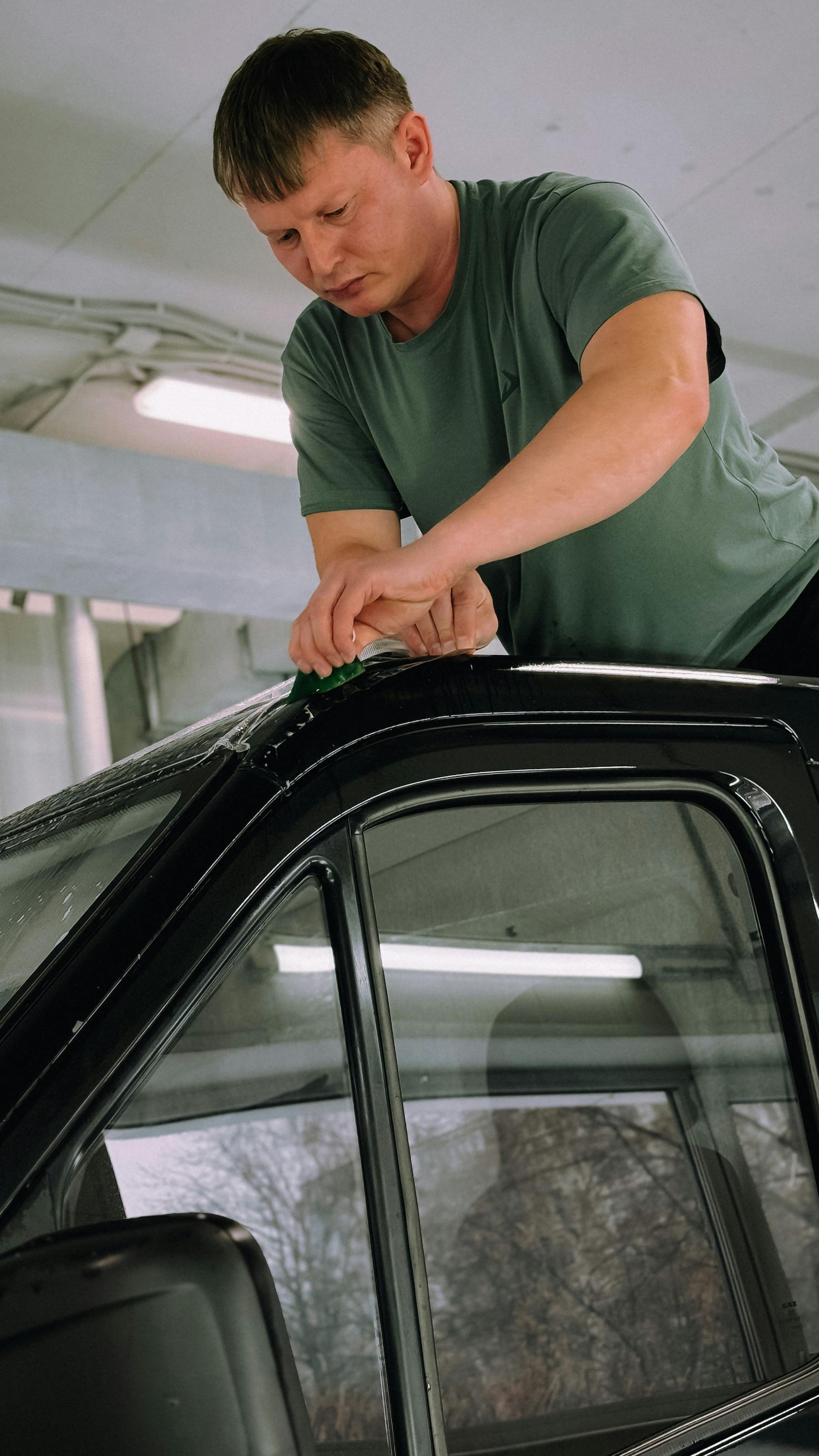 Man in green shirt working on the roof of a black car in a garage.