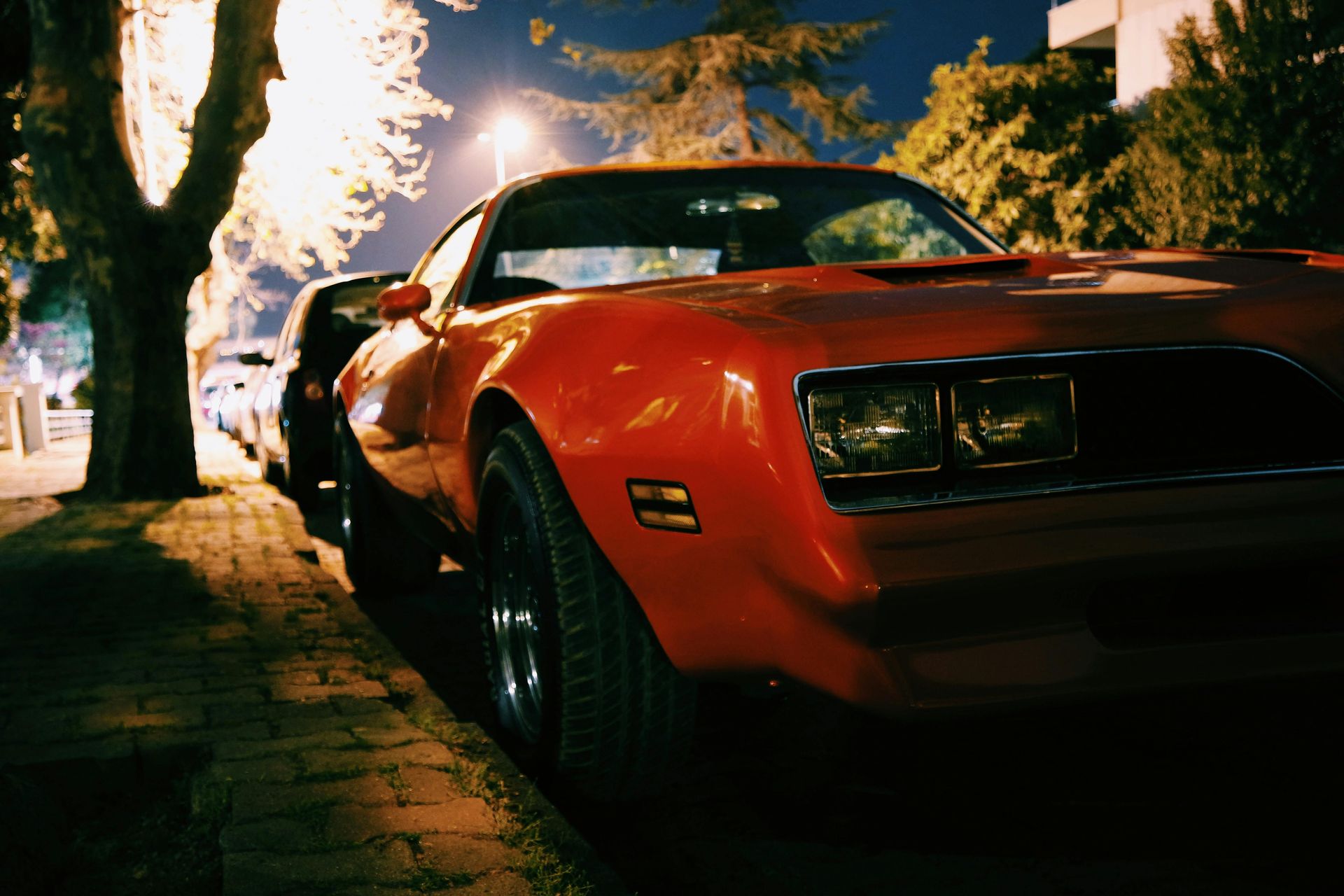 Red classic car parked on a brick-lined street at night, illuminated by streetlights, with a dark sky and trees.