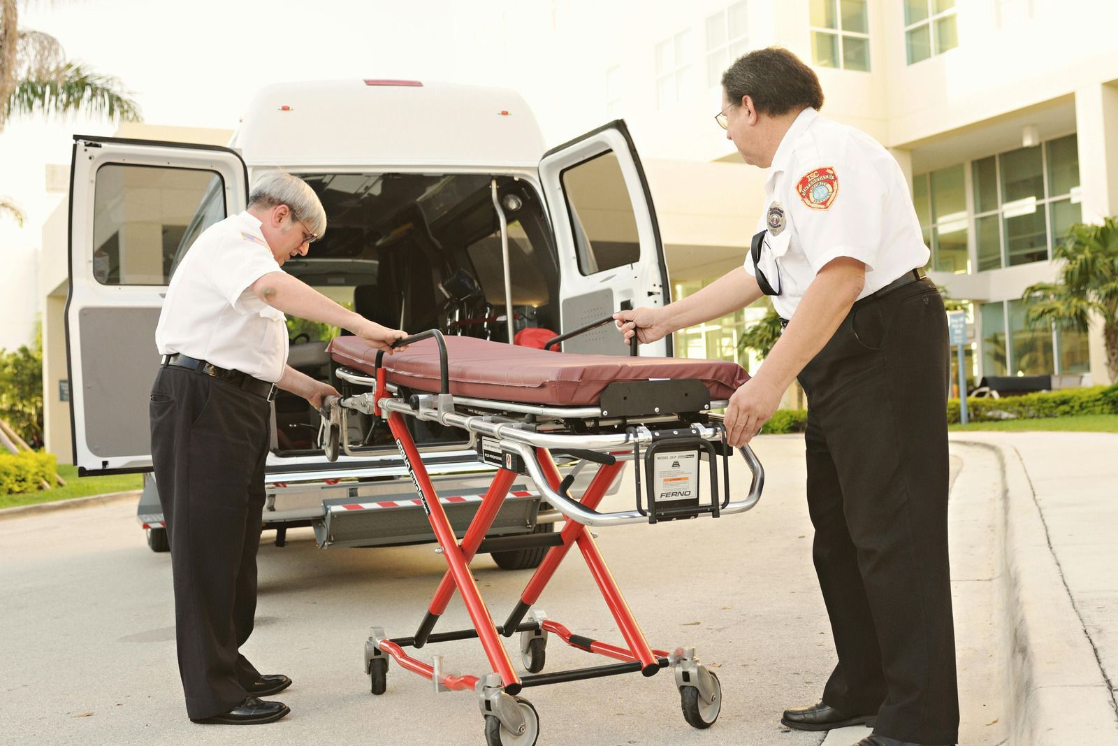 Two paramedics with a red stretcher loading into the back of an ambulance outside a building.