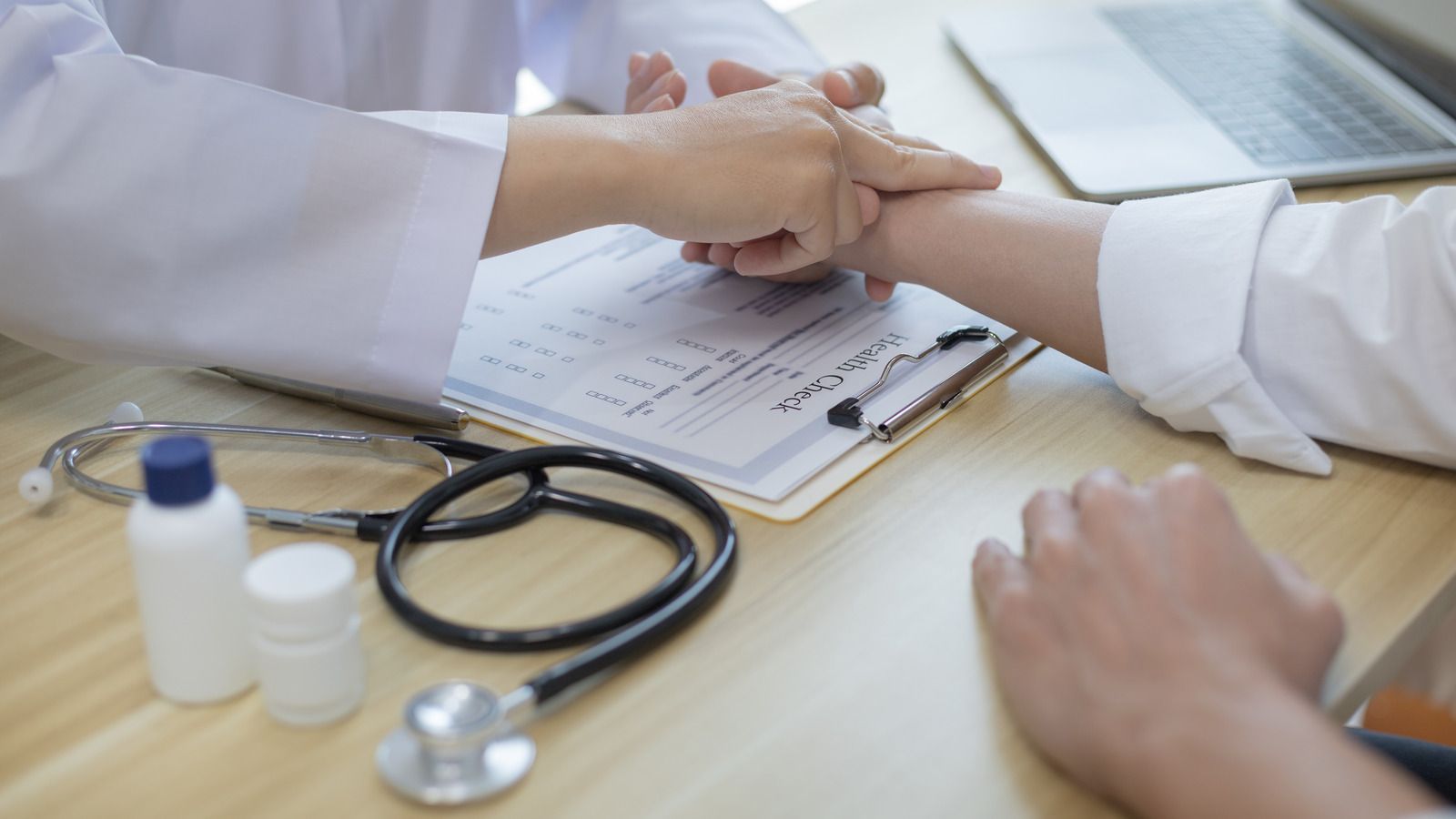 Doctor checking patient's wrist pulse at a desk; stethoscope and medication bottles present.